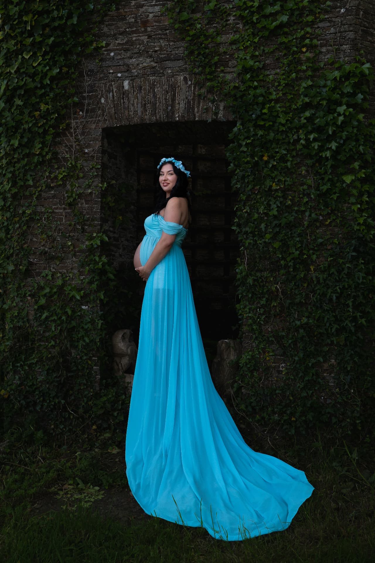 Pregnant woman in a flowing blue dress standing in an ivy-covered stone doorway