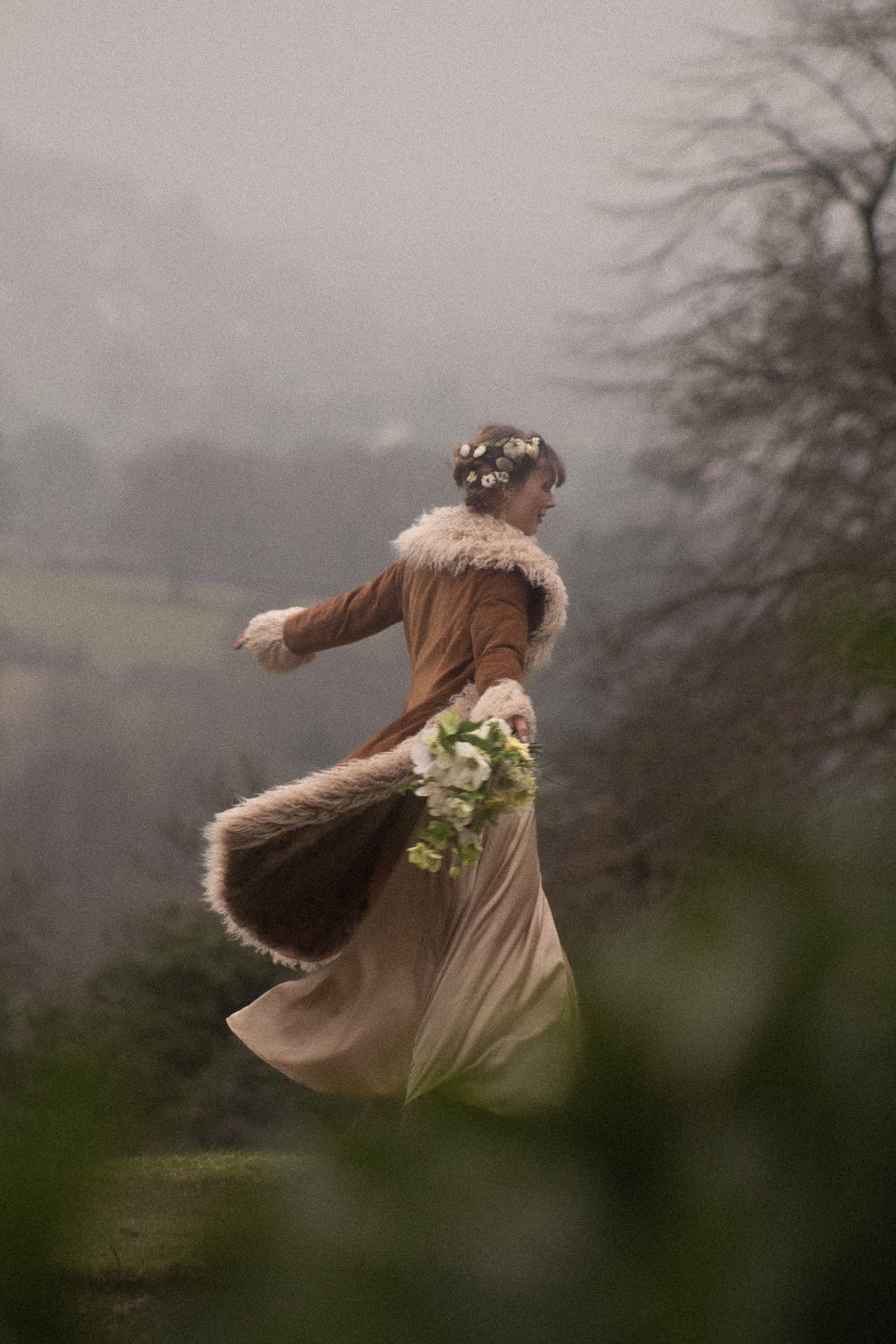 A bride in a fur-trimmed coat with flowers in her hair, twirling in a misty woodland