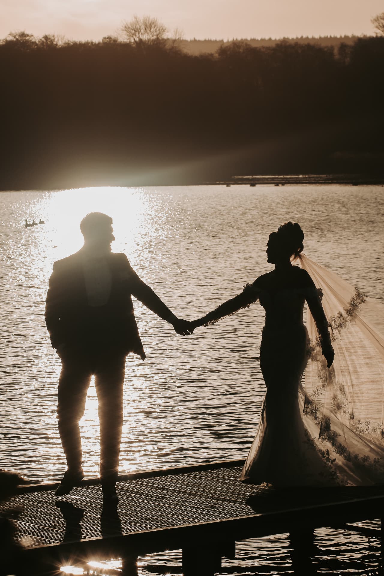 Bride and groom holding hands at sunset, silhouetted against a shimmering lake