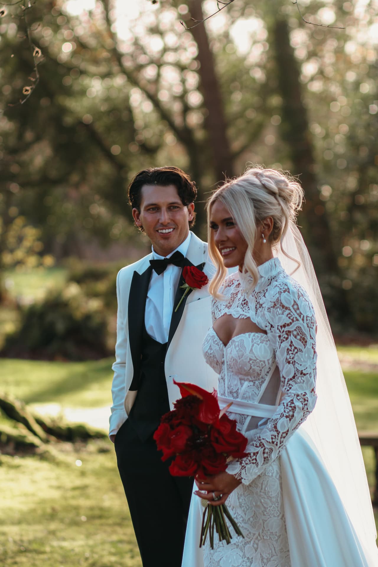 Bride in white lace dress with red bouquet and groom in white tuxedo standing together in a sunlit garden