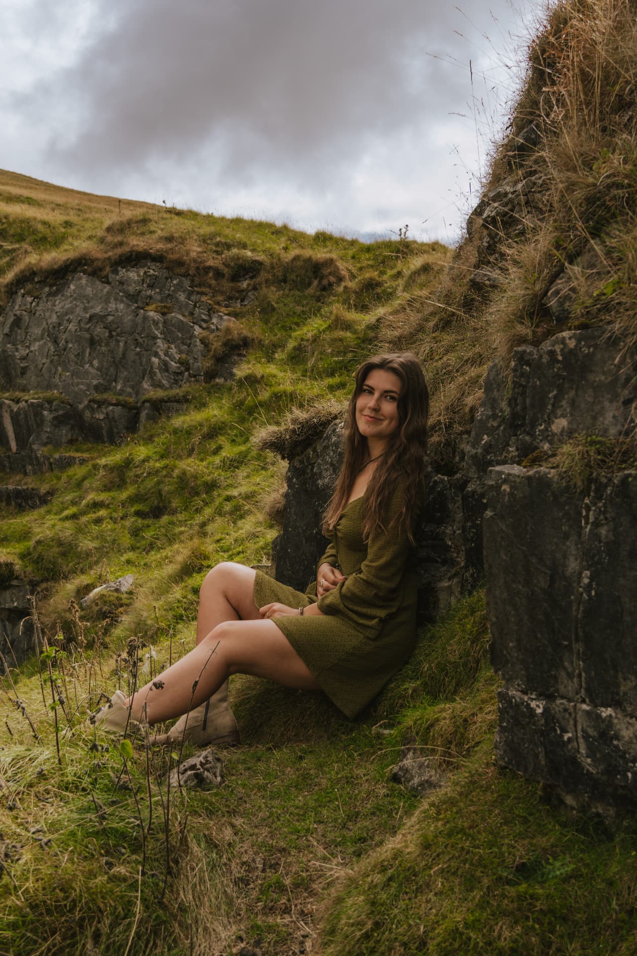 Natalia in an olive green dress sitting among mossy rocks and grass on a mountainside