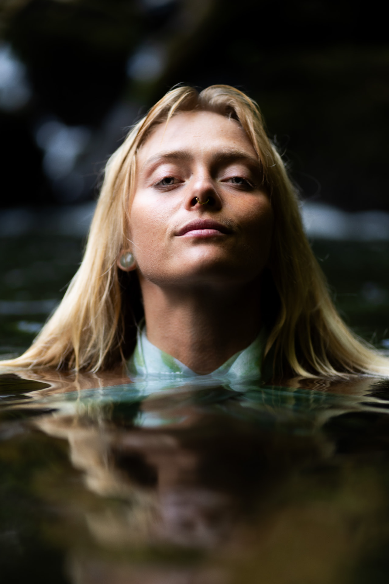 Serene portrait of a blonde woman with a nose ring floating in still water, surrounded by dark reflections