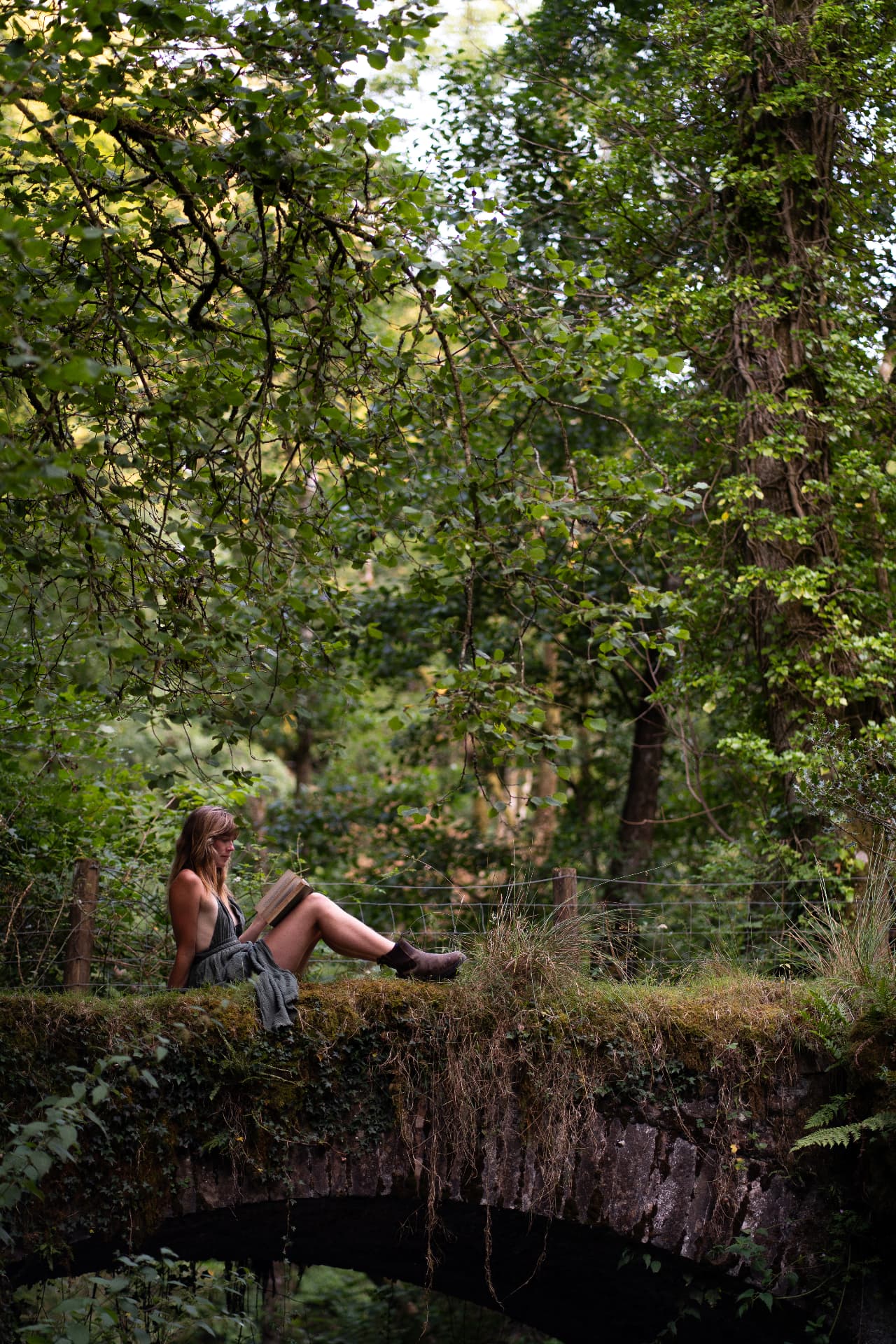 Woman reading a book while sitting on a moss-covered stone bridge in a sunlit woodland