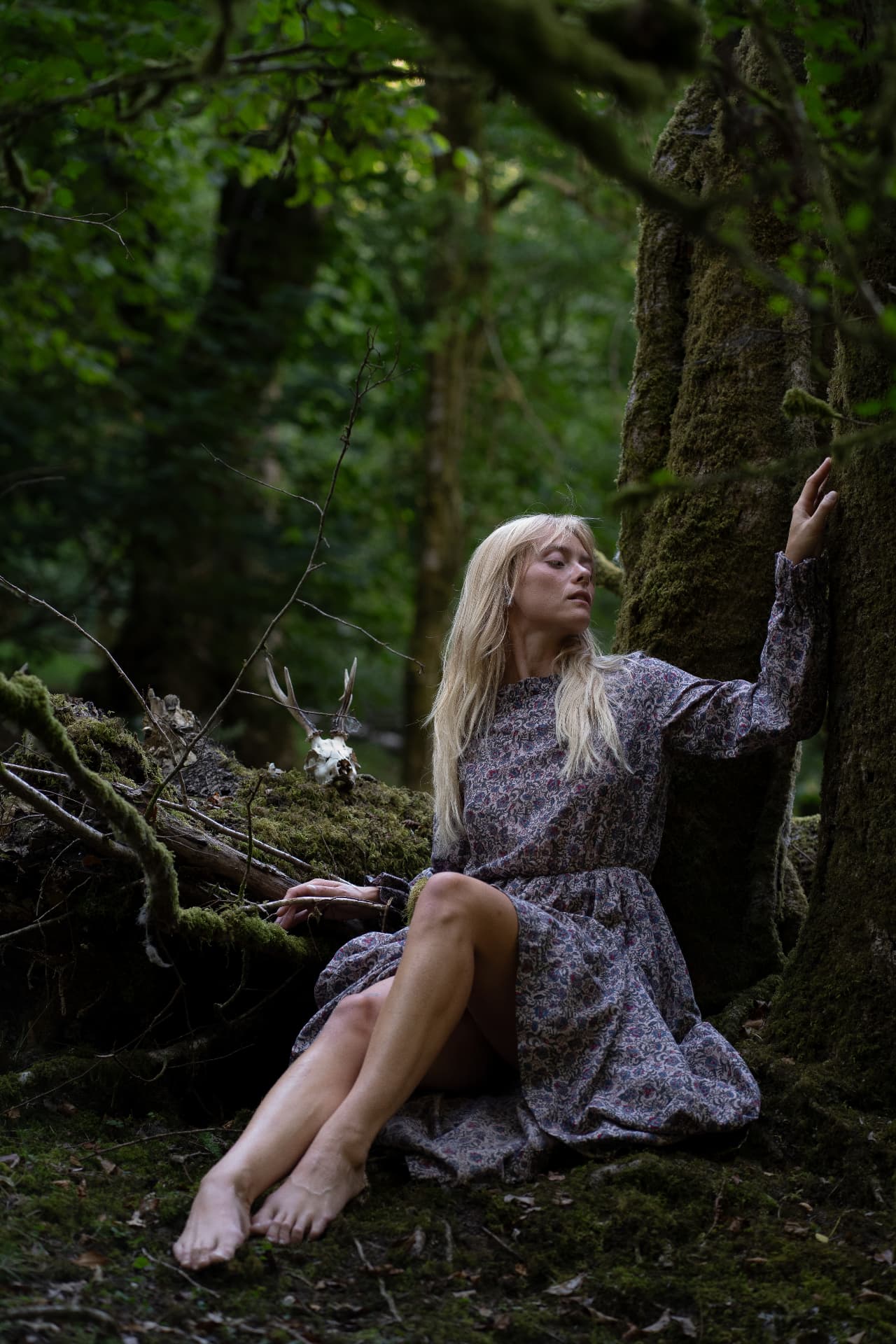 Blonde woman in a floral dress reclining against a moss-covered tree in a dark woodland